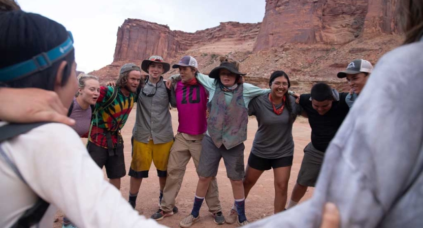 a group of people wrap their arms around each other in front of a vast red rock landscape.